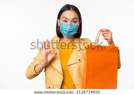 Girl in medical face mask from covid-19, showing shopping bag of store in mall, okay sign, recommending discount in shop, standing over white background