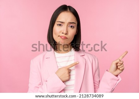 Skeptical asian businesswoman, saleswoman sulking and looking with disapproval while pointing fingers right, showing bad info, upsetting news, standing over pink background