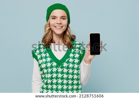 Happy young brunette girl teen student wears checkered green vest hat hold in hand use mobile cell phone with blank screen workspace area isolated on plain pastel light blue background studio portrait