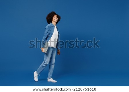 Full body side view fun young smiling happy black woman in casual clothes shirt white t-shirt hold closed laptop pc computer isolated on plain dark blue background studio. People lifestyle concept.