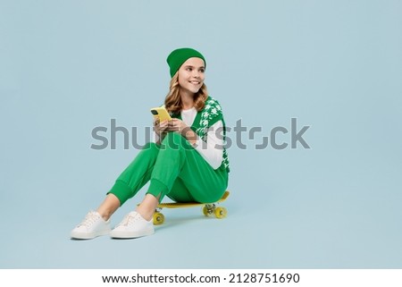 Full size body length cheery young brunette girl teen student wears checkered green vest hat sit on skateboard hold use mobile cell phone isolated on plain pastel light blue background studio portrait