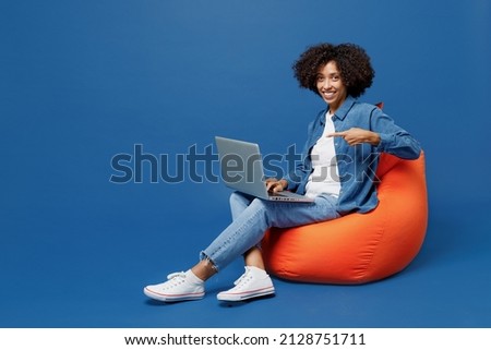 Full body young smiling happy black woman in casual clothes shirt white t-shirt sit in bag chair hold use work poing finger on laptop pc computer isolated on plain dark blue background studio portrait
