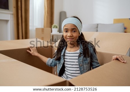 A cute little girl with a hairband sits in a cardboard box used for packing things during a move, sister hides from her brother while they play