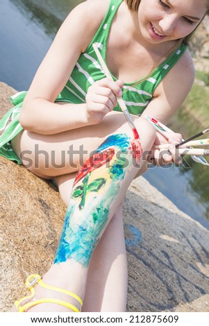Young adult artist full length girl with body art on leg painting an summer landscape near clear water Woman sitting on stone mountain beach Empty copy space for inscription