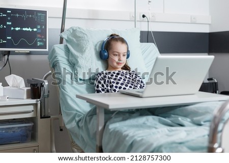 Young patient girl sitting in pediatric hospital bed while watching funny cartoons. Sick little child using computer to watch comical content on internet while sitting in pediatric clinic ward room.