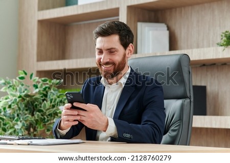 Businessman is smiling and typing a message on his smart phone in office. He is in a suit, workplace in modern and nice office Royalty-Free Stock Photo #2128760279