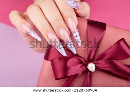 Hand with long artificial french manicured nails decorated with gemstones holding a gift box. 