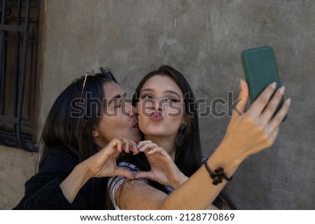 Latin American mother (50) and daughter (24) take a selfie, both make a heart symbol with their hands while their mother kisses her daughter affectionately. Mother's day concept and technology.