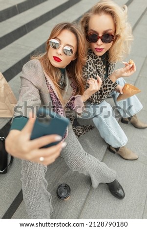 Glamorous young beautiful girls in fashionable clothes sitting on the steps, drinking coffee and taking selfies on their smartphones