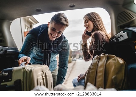 Young beautiful woman talking on the phone making a call from the back of her car sitting in the trunk while her boyfriend or husband it taking luggage baggage and other belongings from the car travel Royalty-Free Stock Photo #2128833014