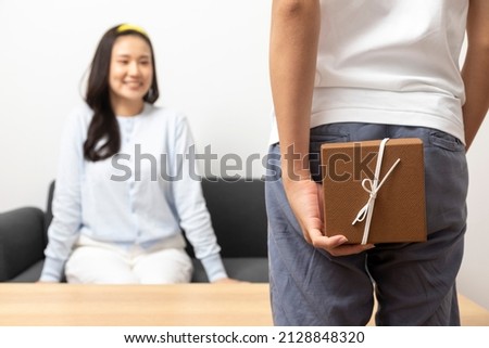 Young asian man hide a gift box with woman sitting on a sofa in living room relax and resting ,happy lover romantic couple concept.