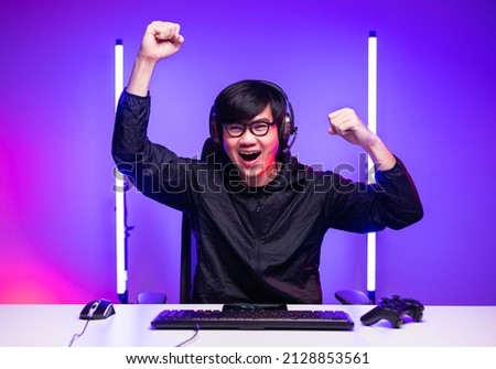 Excited and shocked face of Asian gamer with headphone playing computer pc video game online sitting at living room. Young professional gamer streaming on social playing game very fun
