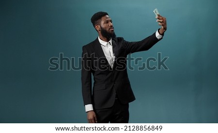 A pretty african man with a dark beard in a white shirt and black suit is taking some photos on his phone