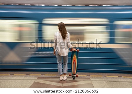 Woman wait for metro car at subway station with train passing by on background. Back rear view of young girl with colorful longboard skate casual student look for underground arrival alone on platform