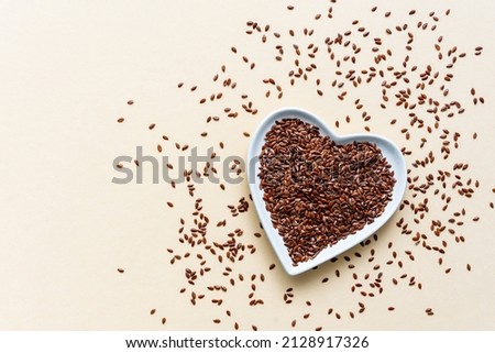 Whole flaxseeds in a white heart shaped bowl on a beige background. Top view, healthy eating.  Royalty-Free Stock Photo #2128917326