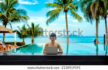 An asian man working on his laptop at. a pool bar with his headphone on, behind him there is the view of the sea, coconut trees and the mountains Royalty-Free Stock Photo #2128919591
