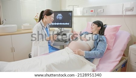 asian pregnant woman lying in hospital ward has uterus utltrasonographic diagnosis - female doctor shows picture on computer and takes notes by digital tablet