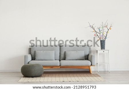Tree branches with Easter eggs in vase on table and sofa near light wall Royalty-Free Stock Photo #2128951793