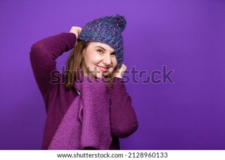 Funny Caucasian Adult Female Woman In Warm Knitted Hat and Violet Scarf Posing with Lifted Hands And Puling Hat Down Against Purple Background. Horizontal Composition