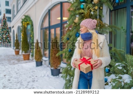 girl with a gift in red jacket and hat standing near a large Christmas tree