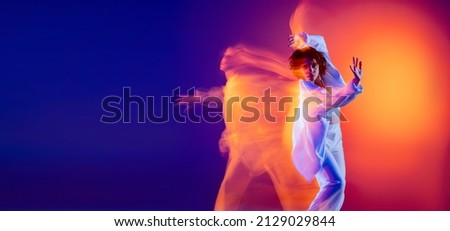 Image of flexible young girl, hip-hop dancer in white outfit dancing hip hop isolated on blue background in yellow neon light. Youth culture, hip-hop, movement, style and fashion, action. Royalty-Free Stock Photo #2129029844