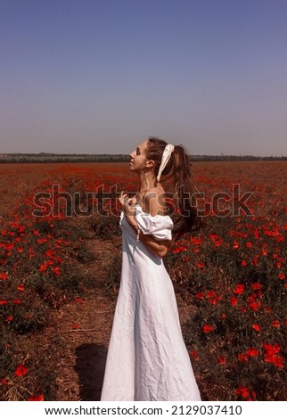 young girl with a bow in the hair in white summer dress is standing in red poppy field and looking away with closed eyes in sunlights on blue sky background. free space