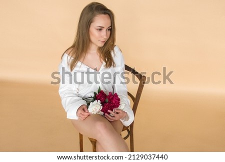 young blonde woman in white shirt is sitting on brown retro chair with red and white peonies in hands and looking away on the beige wall background, free space