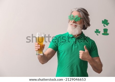 Senior man in eyeglasses with glass of beer showing thumb-up on light background. St. Patrick's Day celebration