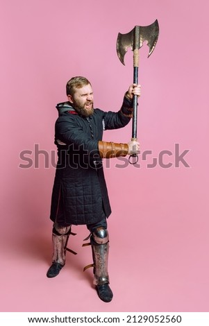 Studio portrait of young bearded man in image of medieval warrior, archery in chain armor with ax isolated over pink background. Ready to fight. Comparison of eras, history, renaissance style