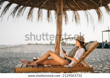 Vacation by sea, weekend on t beach, a beautiful tanned lady in a hat looks at the smartphone screen, sits on a sun lounger on the beach under a straw umbrella. Vacation vacation concept. copy space. Royalty-Free Stock Photo #2129063732