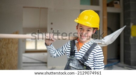 cool boy with yellow safety helmet and dungarees on construction site in a house and loft with shovel in hand