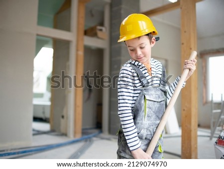 cool boy with yellow safety helmet and dungarees on construction site in a house and loft with shovel in hand