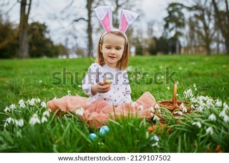 Girl wearing bunny ears playing egg hunt on Easter. Preschooler sitting on the grass with many snowdrop flowers and eating chocolate bunny. Little kid celebrating Easter outdoors in park or forest
