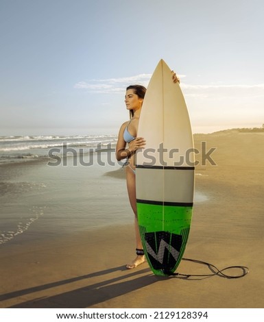 Young beautiful woman standing on the sand looking at the sea with a surfboard in front of her with two in the background. Mention travel, water sports, vacation trips and fun with friends.
