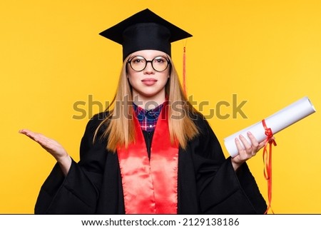 Girl graduate in graduation hat and eyewear with diploma on yellow background. Blonde young woman wearing graduation cap and ceremony robe holding Certificate tied with red ribbon. Education Concept Royalty-Free Stock Photo #2129138186
