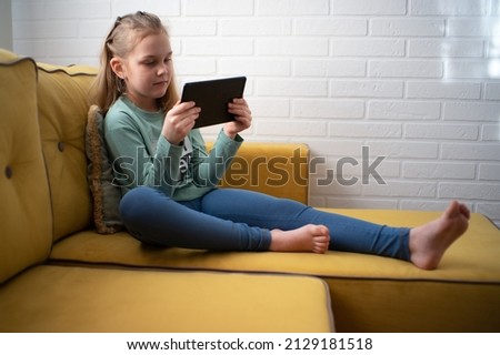  girl is sitting on a sofa and enjoying playing an online game on a digital tablet computer, or listening to an online webinar, or reading a book. Addicted to technology, happy little kid using funny 