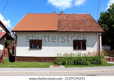 Attached small suburban family house with two owners split in the middle with left half covered with new red roof tiles and right one with dilapidated old roof tiles built next to paved Royalty-Free Stock Photo #2129194241