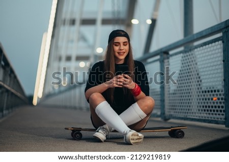 Young woman sitting on her longboard and using a smartphone while on the bridge