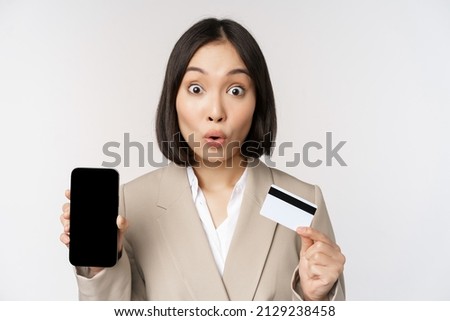 Corporate woman with happy, enthusiastic face, showing credit card and smartphone app screen, standing in suit over white background