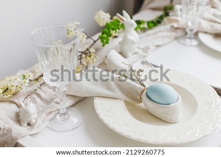 Stylish elegant Easter brunch table setting. Easter egg in bunny napkin on plate with cutlery, bunny, spring flowers on rustic white table. Modern Easter table arrangement Royalty-Free Stock Photo #2129260775
