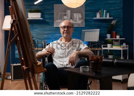 Elderly man sitting in wheelchair drawing black and white sketch using pencil on canvas in artist studio. Senior citizen who is paralyzed feeling creative in front of easel creating fine art.