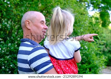 close-up of small child, 2-year-old blonde girl in red dress, old man, grandfather and granddaughter, concept of childhood, family relationships of generations, walk in summer in nature