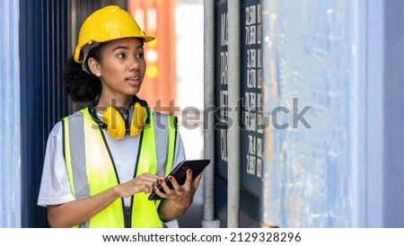 Young African American trainee in safety suit, yellow hard hat and earmuff uses tablet or smart phone in a shipyard. African curly hair female worker in smile. Female loader with yellow earmuff Royalty-Free Stock Photo #2129328296