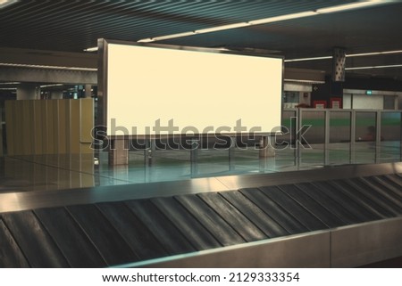A blank advertising poster in an international airport near an empty moving luggage belt. Baggage claim area with a long horizontal advertisement banner or a digital informational billboard mockup