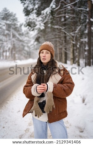 Young millennial asian woman in stylish sheepskin coat and knitted hat looking thoughtfully at camera, standing on empty snow-covered road near forest. Winter wardrobe, fashion trends in wintertime
