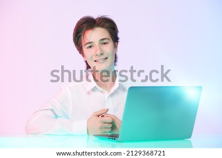 A smart happy teenage boy in a neat white shirt sits in front of a laptop looking at the camera with a satisfied smile. Education. Clever schoolboy. Pink background with red and green light.