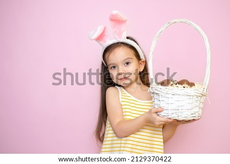 Happy Easter. A cute beautiful girl with bunny ears holds a basket of chocolate eggs in her hands. Pink background. Space for text High quality photo