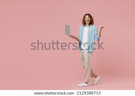 Full length young happy man with long curly hair wear blue shirt white t-shirt hold use work on laptop pc computer do winner gesture isolated on pastel plain pink color wall background studio portrait
