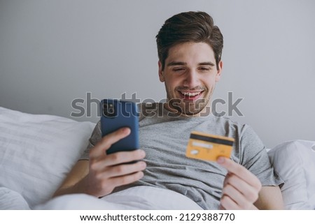Young happy man in pajamas grey sit in bed use mobile cell phone credit bank card for online shop order delivery book tour rest relax at home indoors bedroom Good mood sleeping night bedtime concept