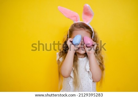 beautiful smiling blonde girl with bunny ears holding an easter egg in her hands, closes eyes, on a yellow background, kid celebrate easter. Royalty-Free Stock Photo #2129395283